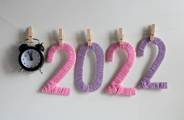 The clock and the numbers 2022 made of pink and lilac threads on a white background hang on clothespins. The concept of the New Year.