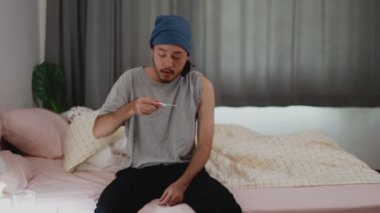 Hasta ve grip olmuş Asyalı bir adam evde yatıyordu. Evde yatak odasındayken termometreyle soğuk hava kontrolü yapan Asyalı bir adam..