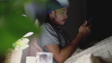 Evde yatarken cep telefonu kullanan Asyalı çekici bir adam. Sosyal medyaya bakmak ve video izlemek.
