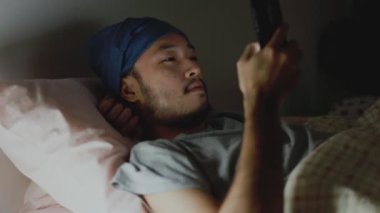 Gece geç saatlerde evde yatarken cep telefonu kullanan yakışıklı Asyalı adam. Sosyal medyaya bakmak ve video izlemek.