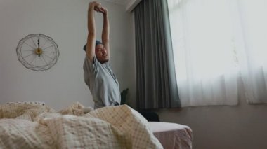 Yakışıklı Asyalı adam yavaşça uyanıyor ve erkenden yataktan kalkıyor perdeleri açmak için ve pencereden dışarı bakıyor. Sabah güneşi evdeki pencereden parlıyor..