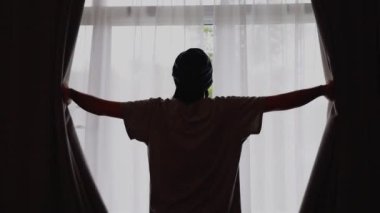 Yakışıklı Asyalı adam sabahın erken saatlerinde perdeleri açar ve pencereden dışarı bakarak gerilir..