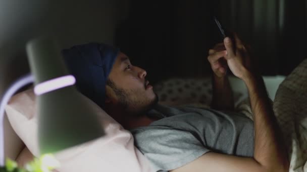 深夜に自宅のベッドで寝そべっている間に携帯電話を使っているハンサムなアジア人男性 ソーシャルメディアの閲覧と動画の視聴 — ストック動画