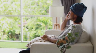 Çekici Asyalı iş adamı koltukta otururken telefonla konuşuyor ve evdeki oturma odasının penceresinden dışarı bakıyor..