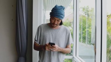 Yakışıklı Asyalı serbest çalışan. Evdeki ofisin penceresinin yanında dikilirken akıllı telefonuna mesaj yazıyor..