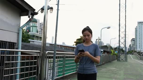 ランニング後にSnsでメッセージを送るスマートフォンを使って歩く魅力的なアジアの女性選手 日没時に路上でワークアウトのライフスタイルをジョギング — ストック動画