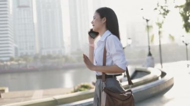 Beyaz tişörtlü Asyalı iş kadını sabah bir parkın önünde akıllı telefon konuşmaları yapıyor..