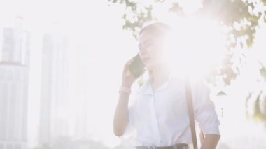 Beyaz tişörtlü çekici Asyalı iş kadını sabah bir parkın önünde akıllı telefon konuşmaları yapıyor..
