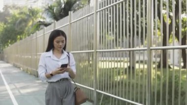 Beyaz tişörtlü çekici bir Asyalı iş kadını akıllı telefon kullanıyor sosyal medyada mesajlaşarak sabah bir parkta yürürken mesajlarını paylaşıyor..