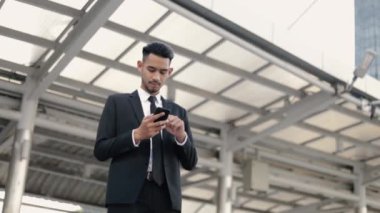 Siyah takım elbiseli genç Asyalı işadamı, büyük bir ofis binasının yakınında güneş ışığı batarken akıllı telefonuyla bir mesaj yazıyor..