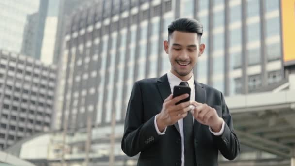 成功したアジアのビジネスマンの勝者は 大規模なオフィスビルの都市の近くの通りに立っている間に幸せなスマートフォンを使用して保持しながら 良いニュースを祝う — ストック動画