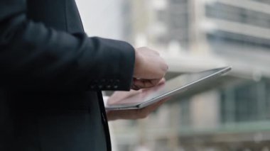 Siyah takım elbiseli Asyalı işadamı, sokakta büyük bir ofis binasının yanında beklerken tabletini kaydırıyor, mesajlaşıyor..