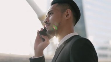 Siyah takım elbiseli Asyalı bir işadamı telefonuyla telefonda konuşurken büyük bir ofis binasının yakınında gün batımını seyrediyor..