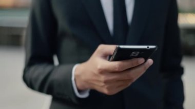 Siyah takım elbiseli yakışıklı Asyalı işadamı akıllı telefonuyla interneti tarıyor, sosyal medyayı süzüyor büyük bir ofis binasının yanında güneş ışığı batarken sokakta dikiliyor..