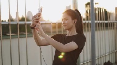 Güzel beyaz kadın atlet akıllı telefon kullanarak koşuyor arkadaşlarıyla selfie çekiyor ve görüntülü sohbet ediyor şehir günbatımında üst geçitte dikilirken.