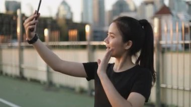 Şehir günbatımında üst geçitte dururken arkadaşlarıyla akıllı telefon paylaşma egzersizi yaparak video sohbeti yapan güzel bir bayan sporcu. Koşu yaşam tarzı sağlıklı.