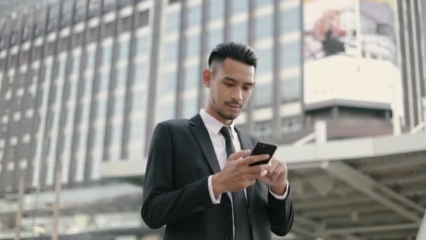 成功したアジアのビジネスマンの勝者は 大規模なオフィスビルの都市の近くの通りに立っている間に幸せなスマートフォンを使用して保持しながら 良いニュースを祝う — ストック動画