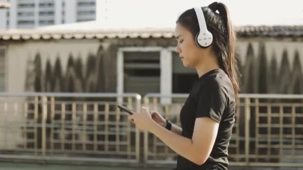 都会を歩きながら音楽を聴きながらスマホを使ってヘッドフォンをして走る幸せな女性アスリート 健康的なジョギングライフスタイル — ストック動画