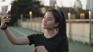 Asyalı bayan sporcular, şehir günbatımında üst geçitte dikilirken arkadaşlarıyla görüntülü konuşma yaparak koşuyorlar. Koşu yaşam tarzı sağlıklı.