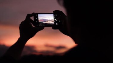 Akıllı telefon kullanan Asyalı adam deniz sahilinde fotoğraf çekiyor. Güzel yaz günbatımı, altın saat ışığı..