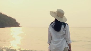 Sahilde durup deniz manzaralı güzel gün batımını izleyen fötr şapka takan çekici Asyalı kadın portresi. Yaz seyahati konsepti.