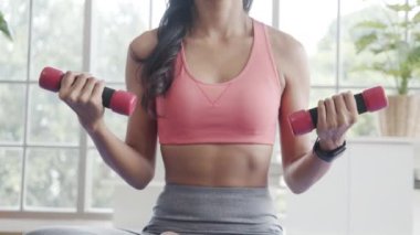 Spor giyim sektöründe aktif Asyalı bir kadın evdeki oturma odasında halterlerle egzersiz yapıyor..