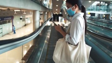 Asyalı kadın turistler cerrahi maske koruması takıyor ve havaalanı terminalindeki yürüyen merdivenlerde akıllı telefonlar kullanıyor ve COVID-19 salgını sırasında sosyal mesafeyi koruyorlar..