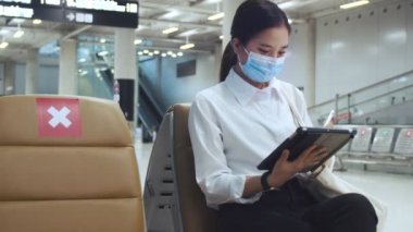 Cerrahi maske takan kadın koruma altında ve havaalanı terminalinde bir sandalyede otururken tablet kullanıyor ve COVID-19 salgını sırasında sosyal mesafeyi koruyor..