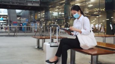Cerrahi maske takan çekici bir iş kadını havaalanı terminalinde bir sandalyede otururken ve COVID-19 salgını sırasında sosyal mesafeyi korurken bir tablet kontrol e-postası üzerinde çalışıyor..