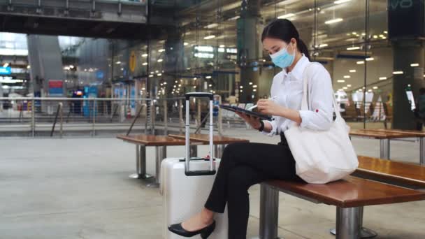 空港ターミナルの椅子に座り Covid 19パンデミックの間に社会的距離を保ちながら タブレットチェックメールで作業外科マスクを身に着けている魅力的なビジネス女性 — ストック動画