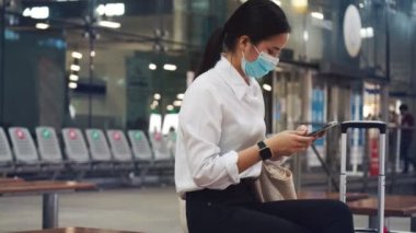 Havaalanı terminalinde bir sandalyede otururken ve COVID-19 salgını sırasında sosyal mesafeyi korurken cerrahi maske takan ve cep telefonu kullanan güzel bir iş kadını..