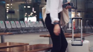 Cerrahi maske takan güzel bir kadın havaalanı terminalinin önünde otururken akıllı telefon kullanıyor ve COVID-19 salgını sırasında sosyal mesafeyi koruyor..