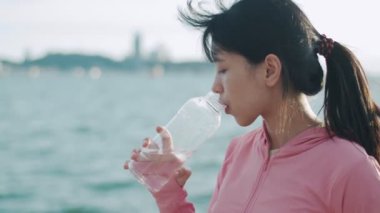 Sahilde dururken su içen Asyalı kadın koşucuya yaklaş. Spor elbiseli bir kız, gün batımını seyrederek spor yapmaya ara veriyor..