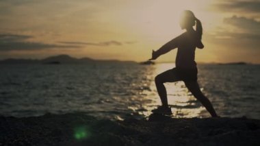 Gün batımında sahilde koşucu esneme egzersizi yapan Asyalı kadın silueti. Güzel bir günbatımında spor giyimli bir bayan sporcu dışarıda koşuyor..