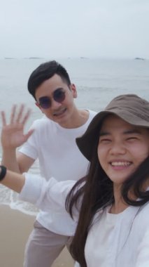 Dikey video: Kafkas çifti sahilde ayakta dururken arkadaş ve aile görüntülerini selamlıyor, yaz tatilinde tatilde olan çift tatilde mutlu..