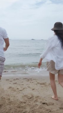 Dikey Video: Asyalı aşık çift koşuyor ve tatilde kollarını havaya kaldırarak başarıyı kutluyor. Plaj gezisinde romantik bir çift..