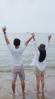 Dikey Video: Asyalı aşık çift hafta sonu tatilinde kollarını havaya kaldırarak başarıyı kutluyor. Plaj gezisinde romantik bir çift..