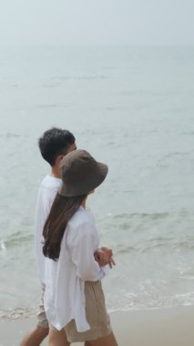 Dikey Video: Mutlu Asyalı çift deniz kenarında yürüyor ve tatilin tadını çıkarıyor, mutlu bir şekilde konuşuyor, gülüyor ve sahildeki manzarayı seyrediyor.