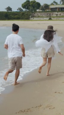 Dikey Video: Genç Asyalı çift el ele tutuşuyor ve tatil haftasonunda kumsalda neşeli bir şekilde koşuyor, Kafkasyalı çift mutlu gülümseyen romantizm deniz kenarında eğleniyor.