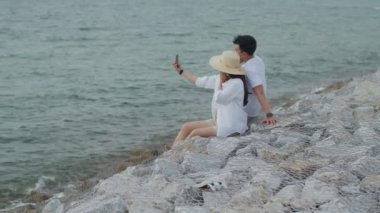 Akıllı telefon kullanan mutlu Asyalı çift, sabah güneşinde deniz kenarındaki taşın üzerinde otururken fotoğraf çektirip selfie çekiyorlar. Romantik çift hafta sonu tatiline çıkıyor..