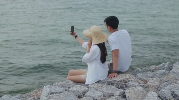 美しい日没の間に海辺の石の上に座っている間 スマートフォンを使用して幸せなカップルは写真と自撮りを取ります 休日の週末にロマンチックなカップル旅行 — ストック動画