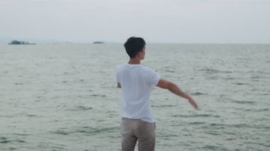 Mutlu Asyalı erkek kutlamaları, hafta sonu kollarını havaya kaldırarak başarıyla kutlanıyor. Yakışıklı beyaz tenli gezgin sahilde duruyor ve deniz manzarası seyrediyor..