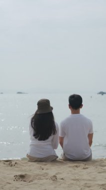 Dikey Video: Asyalı aşık çift tatilde sahilde mutlu mesut otururken, Kafkasyalı çift deniz kenarında mutlu bir şekilde gülümsüyor ve eğleniyor..