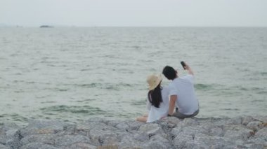 Akıllı telefon kullanan mutlu Asyalı çift, sabah güneşinde deniz kenarındaki taşın üzerinde otururken fotoğraf çektirip selfie çekiyorlar. Romantik çift hafta sonu tatiline çıkıyor..