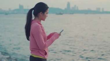 Deniz kenarında koştuktan sonra dinlenme sırasında akıllı telefon kullanarak sosyal medya yayınlarını kontrol eden bir kadın portresi. Spor kadınları ayakta dinlenirken arkadaşlarına mesaj göndermek için cep telefonu kullanırlar..