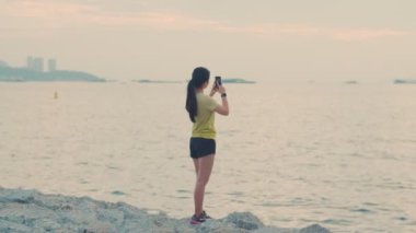 Akıllı telefon kullanan beyaz kadın sporları, güzel bir gün batımında sahilde antreman yaptıktan sonra okyanus manzarası çeker..
