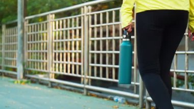 Sporcu kadın elinde su şişesi tutarak koşmadan önce caddede çit kenarında yürüyordu..