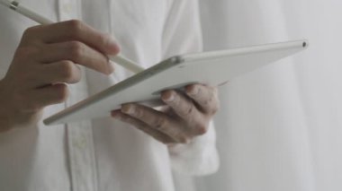 Yakın plan Asyalı işadamı, bir tablet üzerinde stilus kalem kullanarak dijital kontrat imzalıyor ve ofisteki pencerenin yanında dururken tabletin çalışmalarını kontrol ediyor..