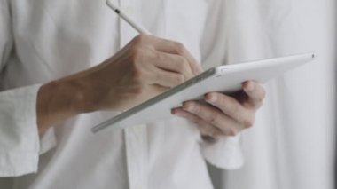 Yakın plan iş adamı, bir tablet üzerinde stilus kalem kullanarak dijital kontrat imzalıyor ve ofisin penceresinin yanında dururken tabletten çalışmaları kontrol ediyor..