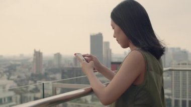 Asyalı bir kadın, günbatımında apartmanın çatısındaki balkonun yanında durup arkadaşlarıyla akıllı telefonlar kullanarak sohbet ediyor..